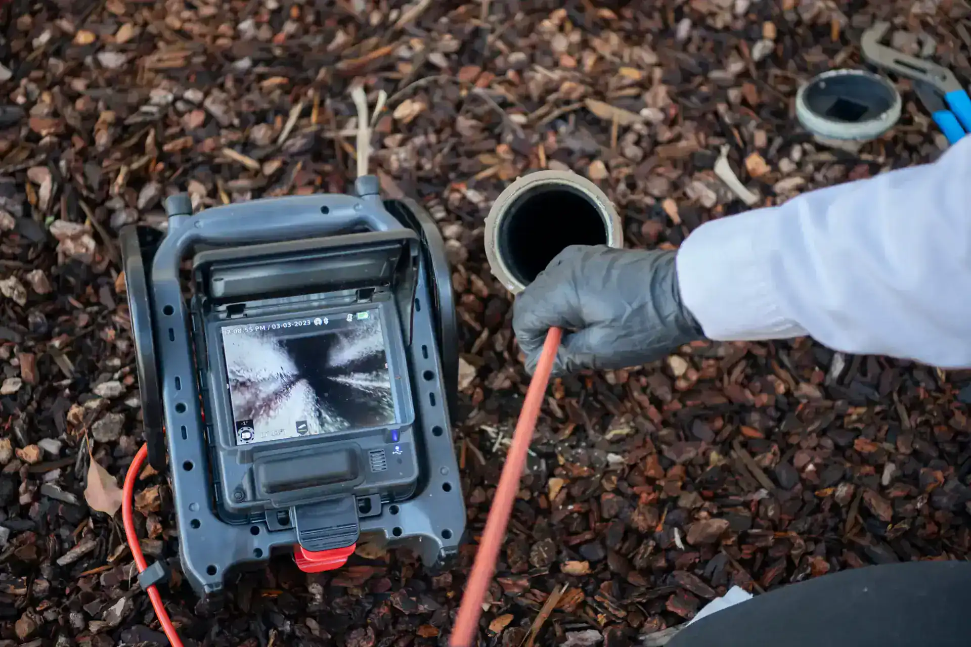 A plumber Pasco County, FL, wearing a glove inserts an inspection camera cable into an outdoor pipe. The camera&rsquo;s monitor displays the pipe&rsquo;s interior, while tools and bark mulch are visible on the ground nearby.