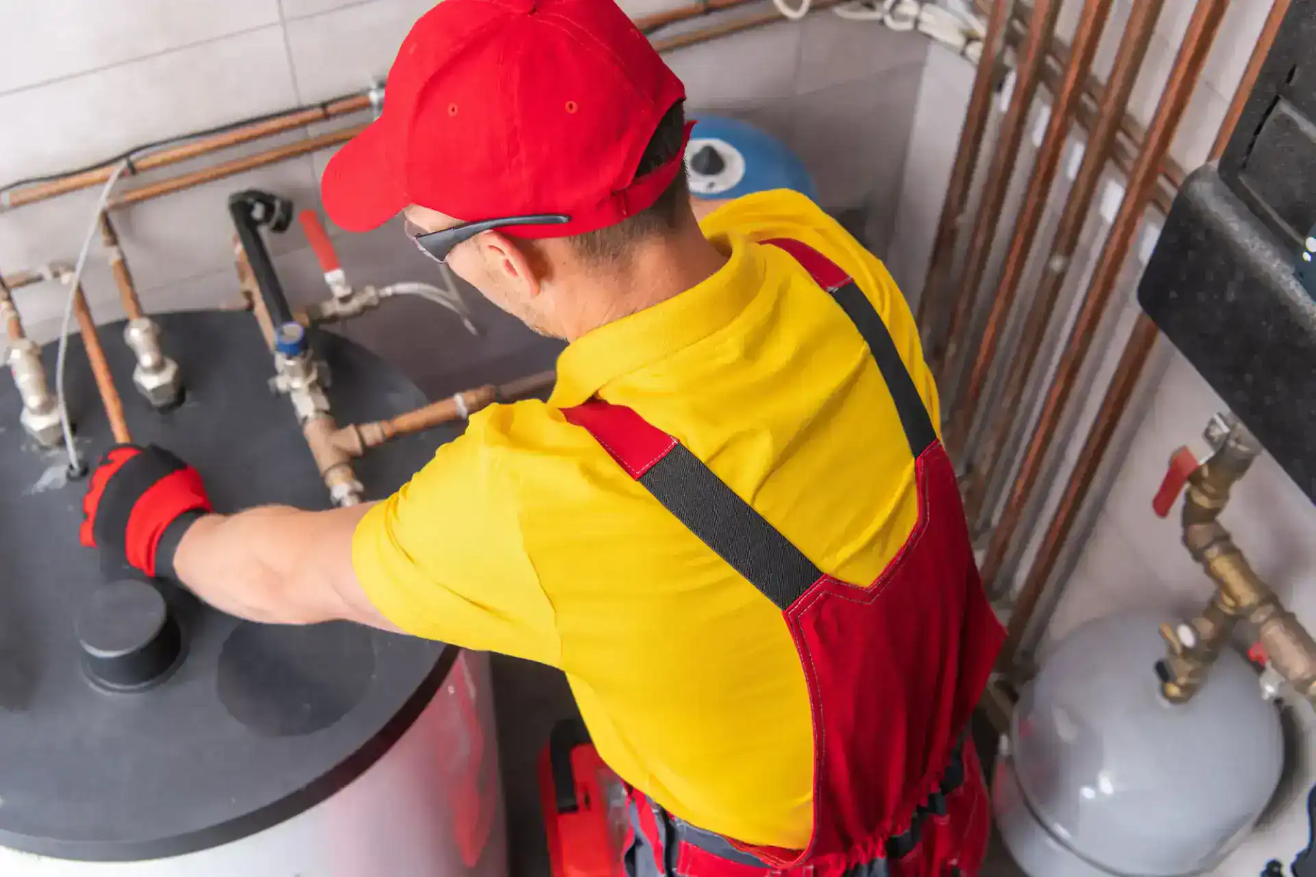A worker in a yellow shirt and red overalls adjusts pipes connected to a water heater in a utility room, surrounded by valves and plumbing.
