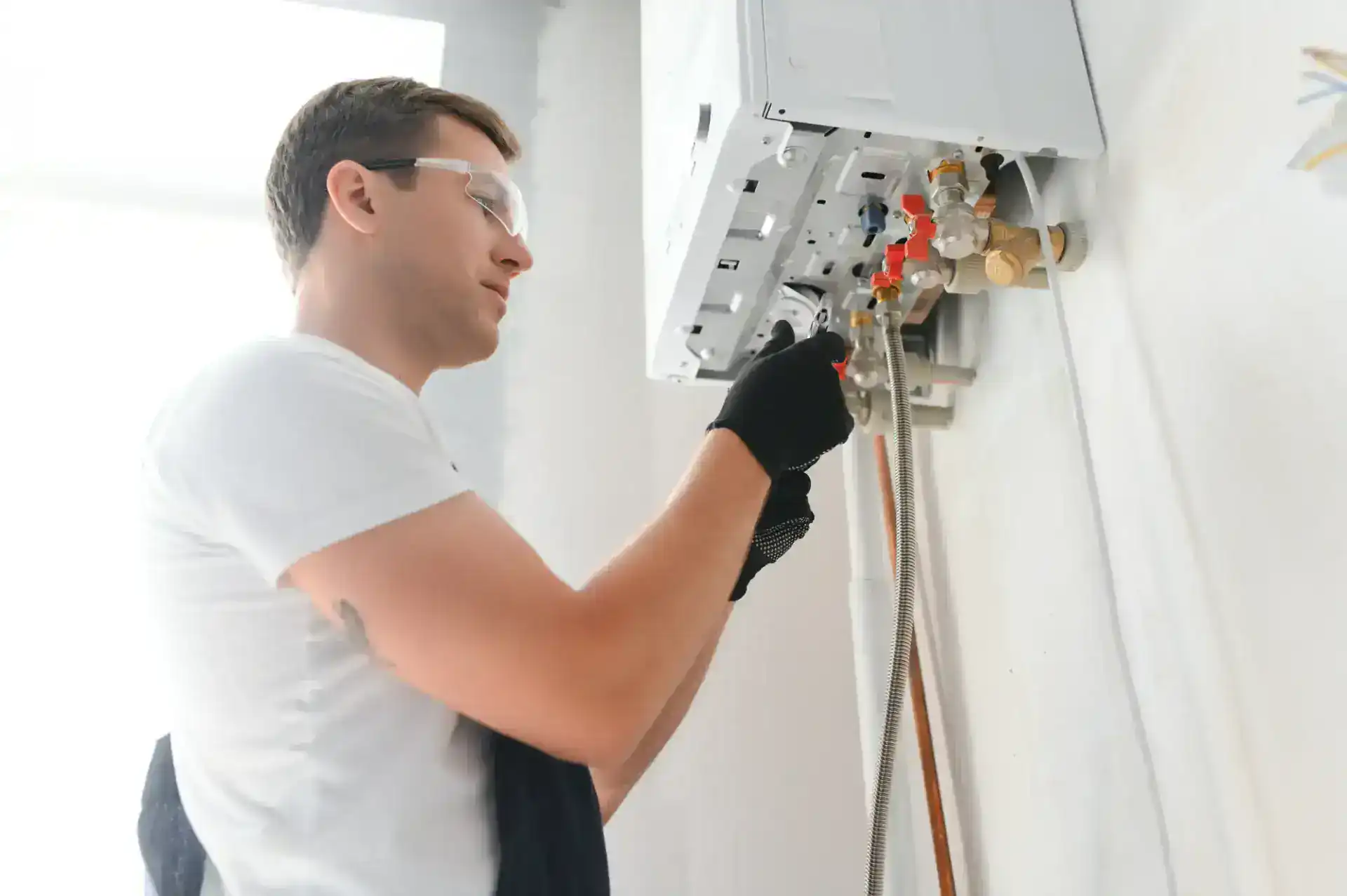 A person wearing safety glasses and gloves adjusts pipes and valves on a wall-mounted boiler or water heater in a bright room.