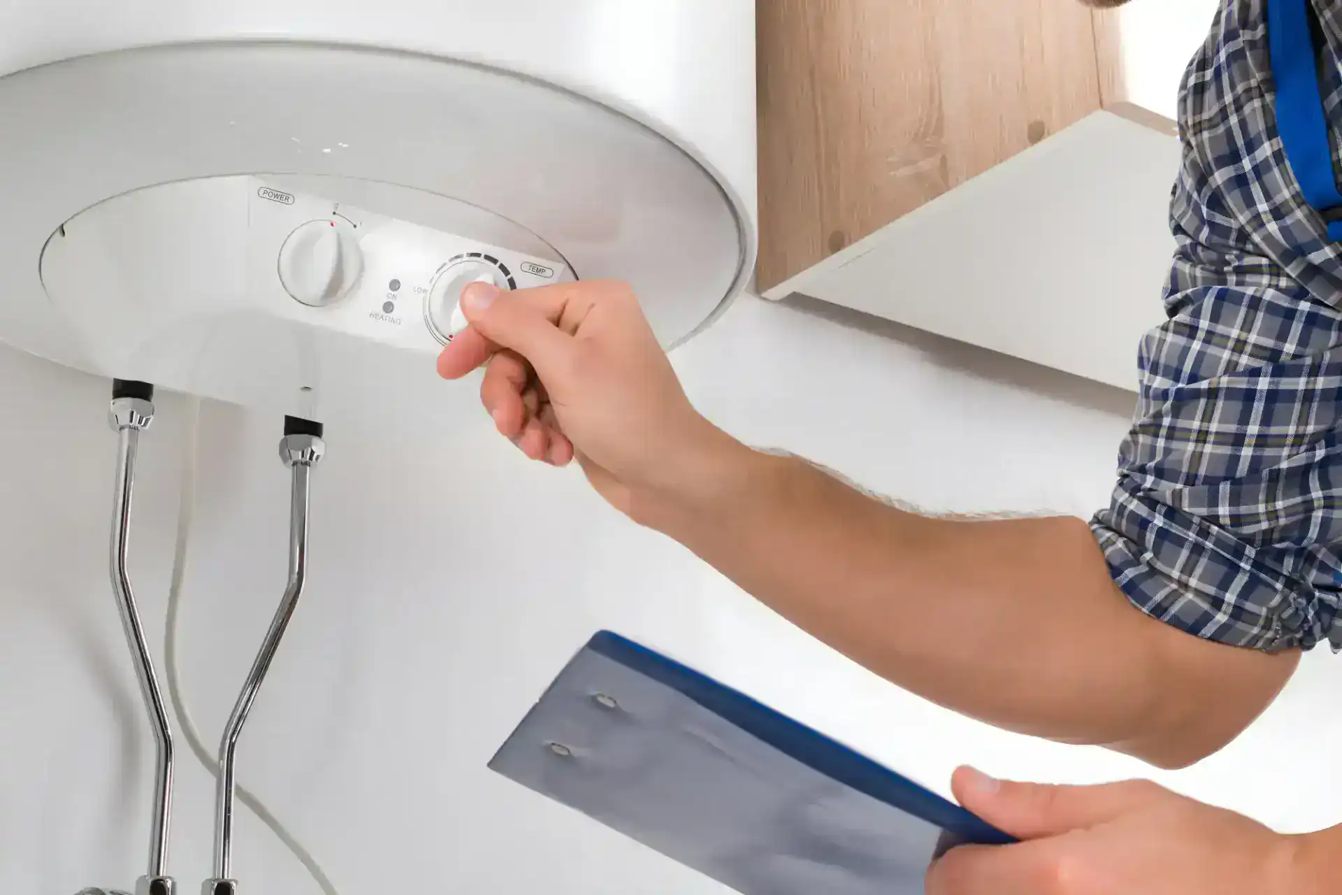 A person adjusts the temperature dial on a wall-mounted water heater while holding a clipboard, with visible plumbing and part of a wooden cabinet nearby.